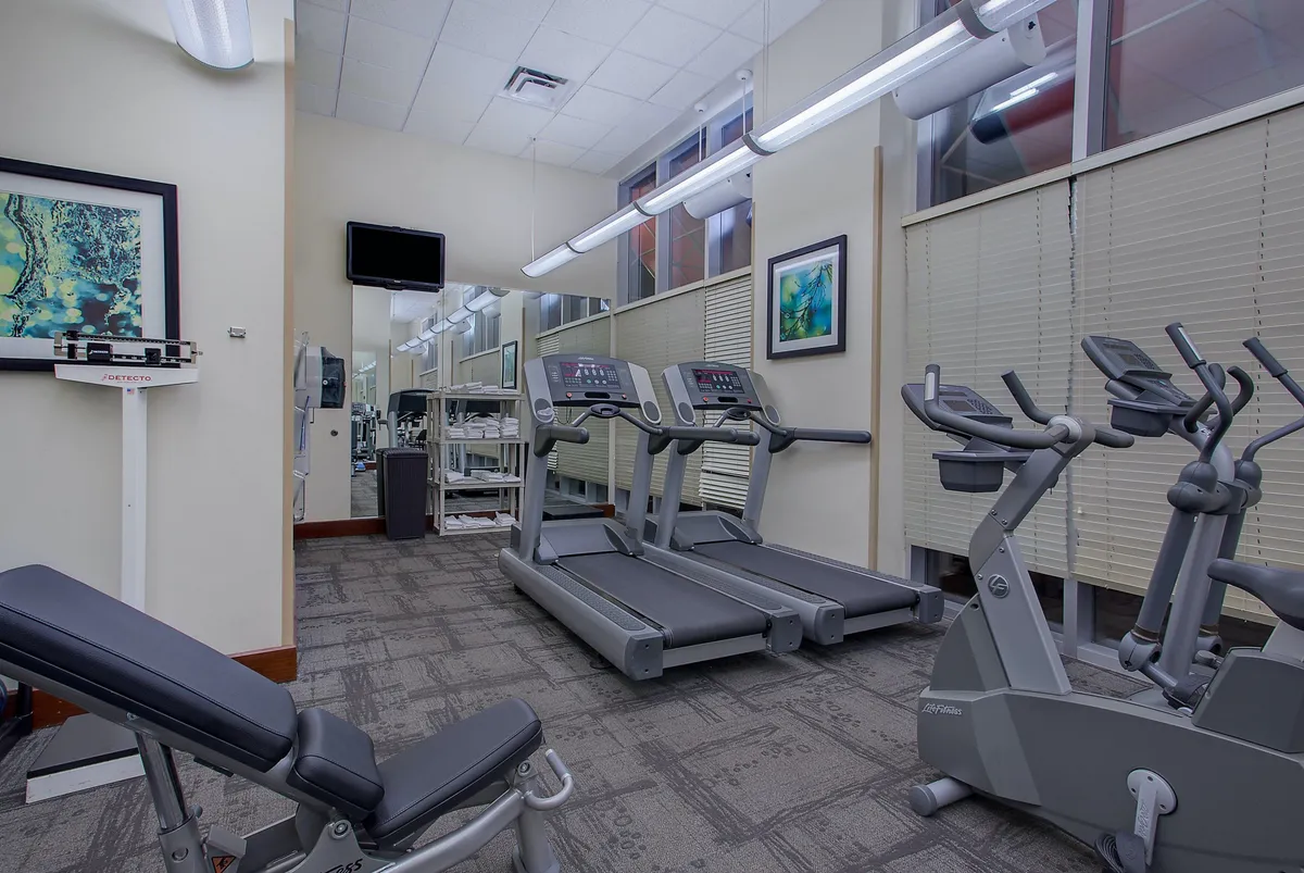 A modern fitness center equipped with treadmills, an elliptical machine, a weight bench, and large mirrors. The energetic mood is set by bright lighting and a sense of activity, with guests seen engaging in personal or group workouts.