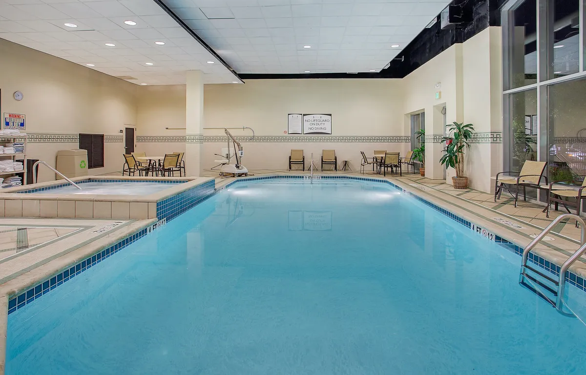 A calming interior scene featuring the shared swimming pool, adjacent lounge chairs, a jacuzzi, and decorative potted plants. The space is bathed in gentle, refreshing light, with visitors enjoying both swimming and quiet poolside moments, encapsulating a leisurely community vibe.