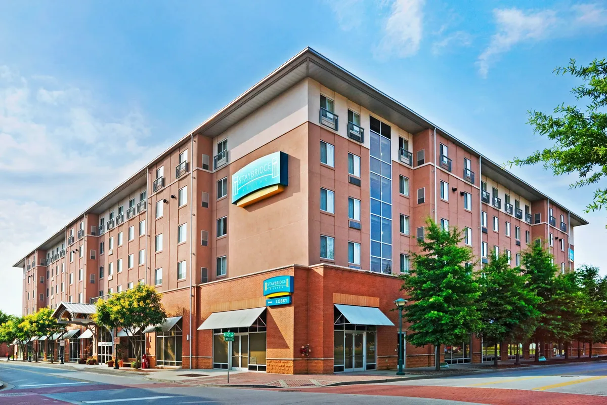 A vibrant exterior view featuring the hotel's modern architecture, with an inviting entrance lobby surrounded by landscaped trees. Guests can be seen strolling through the lively neighborhood, enjoying nearby shops and restaurants, which hints at the walkable downtown location.