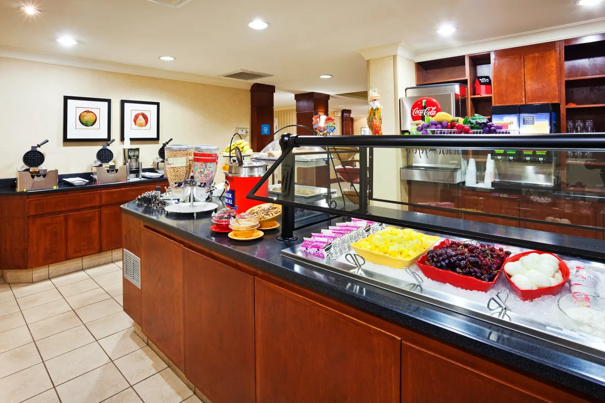A bright, inviting breakfast bar featuring a colorful fruit display, waffle maker, and a well-organized beverage station. The room is bustling with cheerful energy as guests help themselves to complimentary breakfast items, sparking casual conversations and shared morning rituals.