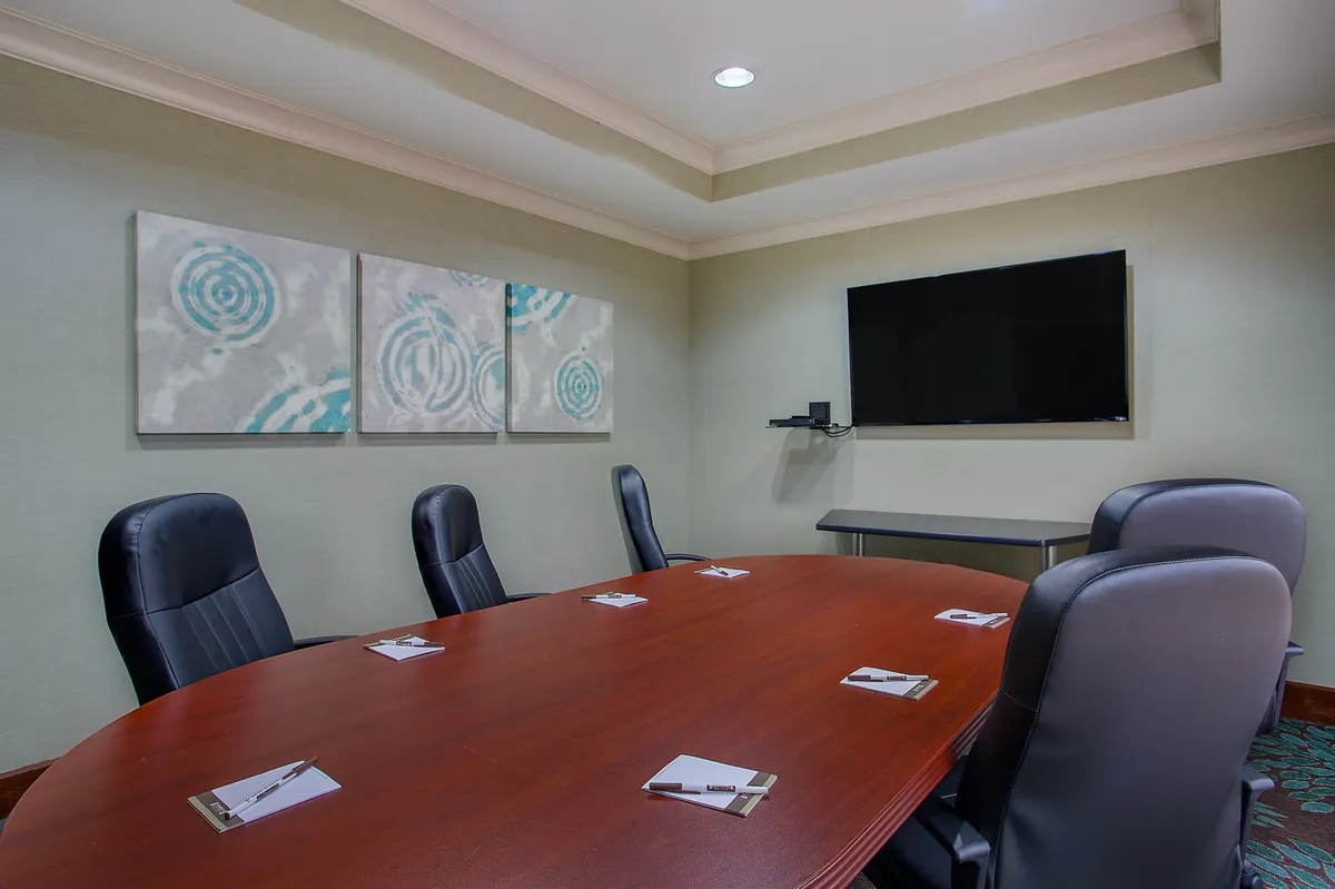 An expansive, well-lit meeting room featuring a large wooden conference table surrounded by high-back leather chairs, with abstract art on the wall and a flat-screen television for presentations. The space is designed for collaboration and formal business meetings.
