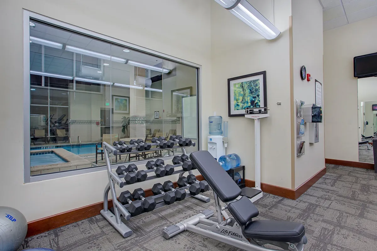 A vibrant fitness center equipped with a dumbbell rack, exercise bench, water cooler, and motivational artwork. The space is energetic and well-lit, with residents engaging in fitness routines, including weightlifting and group exercises. The modern equipment and thoughtful layout create an inviting atmosphere for both solo workouts and social interaction.